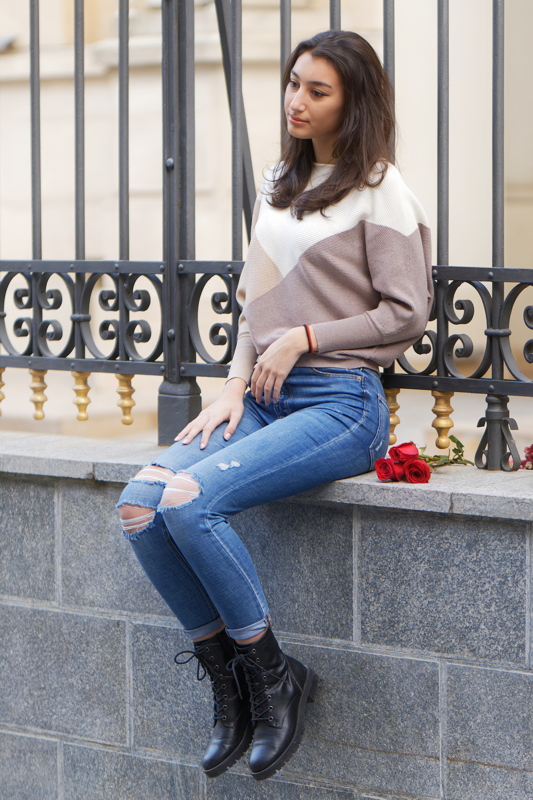 5 - A woman wearing a color-blocked knit sweater, distressed skinny jeans, and black lace-up boots while sitting outdoors near an iron fence